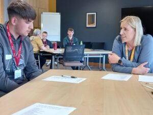 Two people sitting at a table having a discussion during a training or workshop session, with other participants working at tables in the background.