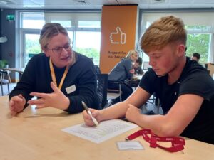 Two people sitting at a table in a training session, discussing a document while one person writes notes, with others working in the background.