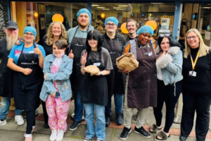 A group of people standing outside a bakery wearing aprons and hair nets, smiling together and holding bags of freshly baked bread.