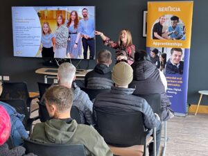 A presenter speaking to a small audience during an apprenticeships information session, with a presentation screen and Bernicia apprenticeships banner in the room.