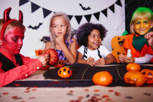 A group of children wearing Halloween costumes sit around a table decorating small pumpkins, with Halloween bunting and pumpkin decorations in the background.