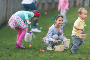 Several children search for colourful eggs on a grassy area, holding small baskets while taking part in an outdoor egg hunt.