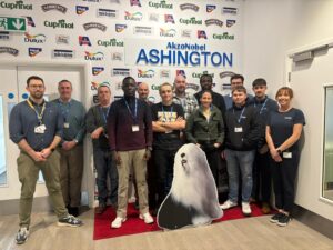 A group photo of Bernicia, AkzoNobel and course participants standing together at the AkzoNobel Ashington site, with branded logos behind them and a Dulux dog display at the front.